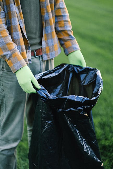 The image depicts a person outdoors holding a large black plastic rubbish bag that is partially open, revealing an empty interior. The individual is wearing light green rubber gloves, a green shirt, a